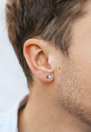 Silberner Steckerohrring im durchstochenen Ohrläppchen, zylindrische Form, glatte Textur, dezenten Glanz, minimalistisches Design, Nahaufnahme von Ohr und Haar.