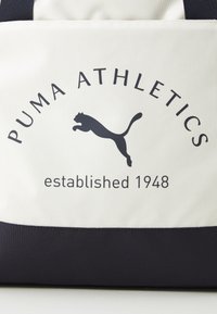 Witte stoffen tas met zwarte accenten, voorzien van de tekst "PUMA ATHLETICS" en een springende Puma-logo. De tekst luidt "opgericht 1948."