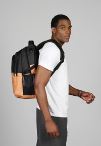 Mochila negra y naranja con bolsillo de malla, que presenta dos compartimentos con cremallera y correas ajustables, llevada en el hombro derecho.