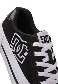 Svart sneaker med en ovandel av canvas, vita snörning, framträdande broderad logotyp, slät svart hälakcent och vit gummisula med svart rand.