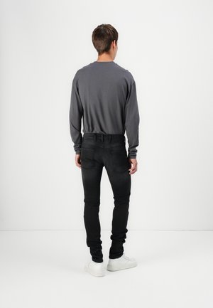 Homme debout face à un mur blanc, portant une chemise à manches longues gris foncé, un jean noir et des baskets blanches.