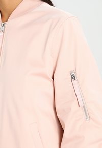 Giacca bomber rosa chiaro con una texture liscia, colletto a coste, zip frontale e tasca con zip sul maniche. Design semplice con accenti minimi.
