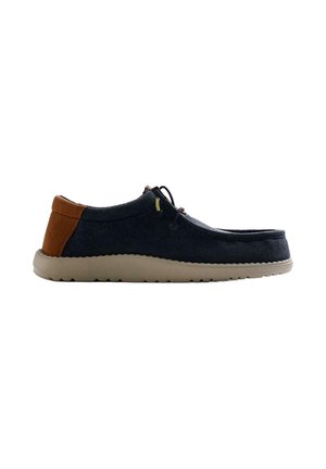 Zapato casual de hombre con cordones, parte superior de tela oscura, parche marrón en el talón y suela de goma beige, visto de perfil sobre fondo blanco.
