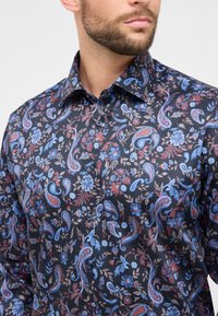 Mann trägt ein dunkles, geknöpftes Hemd mit einem blau-roten floralen Paisleymuster, zeigt Hals, Kragen und Teile des Gesichts.