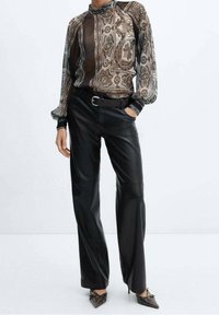 Blusa trasparente a fantasia in marrone e crema, abbinata a pantaloni in pelle nera a vita alta. Ha maniche lunghe e vita con cintura.