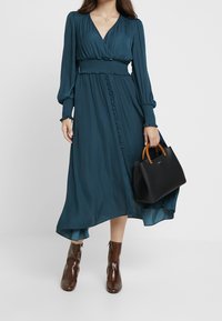 Frau in einem langen türkisfarbenen Kleid mit Knöpfen, braunen Stiefeletten und einer schwarzen Handtasche mit Holzgriffen vor einem einfarbigen Hintergrund.