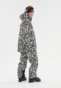 Lumppailuasu, jossa on hupparitakki ja mustavalkoisilla tekstikuoseilla varustetut housut. Mukana hiihtolasit ja kestävät mustat saappaat.