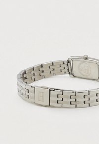 Ezüst rozsdamentes acél karóra négyszögletes számlappal, láncos szíjjal és kapcsos záródással. A hátoldalán gravírozott logó és jelzések láthatók.