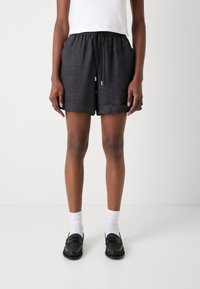 Pantaloni corti neri in lino con vita elasticizzata, due tasche frontali e risvolti. Indossati con una maglietta bianca e mocassini neri.