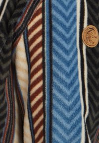 Tejido de forro polar a rayas con patrones en chevron y diagonales en marrón, azul, crema y negro, que presenta un parche redondo de cuero con un diseño en relieve.