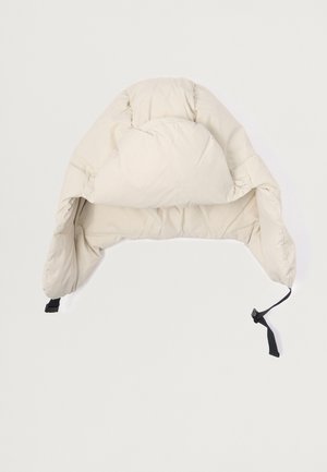 Pehmeä, kermaisen värinen huppu, jossa on pyöreä yläosa, säädettävä musta hihna ja sileä kangas. Ei näkyviä kuvioita tai metalliosia.