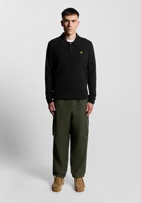 Czarna koszula polo z długim rękawem, kołnierzykiem i małym żółtym emblematem, w połączeniu z oliwkowymi spodniami typu cargo i beżowymi butami.