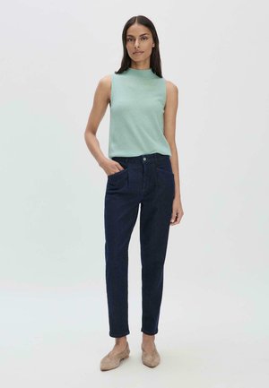Mouwloze mintgroene top met een hoge hals, gecombineerd met donkerblauwe jeans met voorzakken en een getailleerd ontwerp. Beige platte schoenen maken de look compleet.