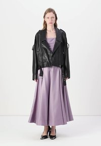 Jaqueta de moto em pele preta, ajustada com fechos e correias, sobre um vestido longo de cetim lavanda fluido, combinado com saltos pretos.