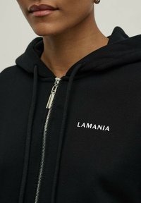 Juodas užtrauktukas hoodis, pagamintas iš minkštos medžiagos, su raišteliu ant kapučio. Ant kairiojo šono yra baltas „LAMANIA“ logotipas ir sidabriniai užtrauktuko elementai.