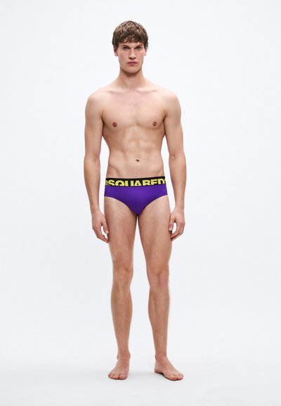 Lila rövidnadrág fekete derékpánttal, amelyen sárga "DSQUARED2" logó található. Sima textúra, testhez simuló kialakítás. A modell egyenesen áll mezítláb.