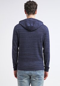 Pull à capuche bleu marine en tissu texturé, présentant un motif à rayures subtil. Poignets et ourlet ajustés, assortis à un jean bleu clair.