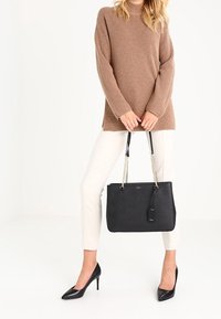 Sac fourre-tout noir à surface texturée, avec des poignées doubles en chaîne et cuir, et une étiquette. Porté avec un pull beige et un pantalon blanc. Design élégant.