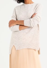 Eine Frau mit verschränkten Armen, die einen weichen, hellgrauen Pullover mit seitlichen Schlitzen und einen beigen, plissierten Rock vor einem weißen Hintergrund trägt.