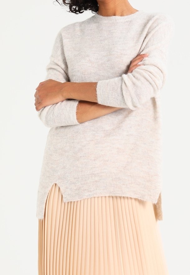 Eine Frau mit verschränkten Armen, die einen weichen, hellgrauen Pullover mit seitlichen Schlitzen und einen beigen, plissierten Rock vor einem weißen Hintergrund trägt.