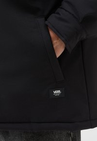 Zwarte Vans-jas met een gestructureerde stof, zichtbare zak en een merklabel op de zoom. Het ontwerp legt de nadruk op functionaliteit.