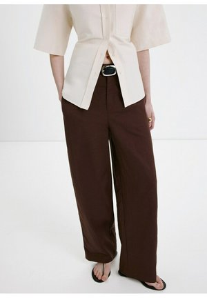 Personne portant un haut crème à manches courtes boutonné, un pantalon large marron taille haute avec ceinture noire, et des sandales noires style tongs sur fond blanc.