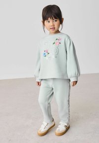 Jasnozielony sweatshirt i dopasowane spodnie z haftem w kwiaty oraz teksturowanym detalem po bokach; zestawione z białymi butami z brązowymi podeszwami.