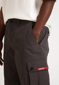 Pantalons cargo en tissu texturé gris foncé, dotés d'une poche latérale avec une étiquette rouge "JACKIT" et une fermeture à bouton. Coupe décontractée avec une taille haute.