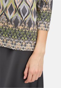 Multicolored patterned top with gray, green, and peach geometric designs; features three-quarter sleeves and a relaxed fit, paired with a dark skirt.
