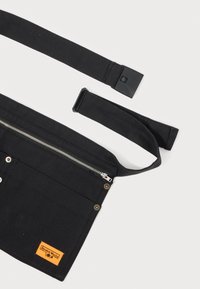 Svart kanvas verktøysbelteveske med glidelåslomme, messingknapper og oransje merkeetikett. Har justerbar stropp for sikker passform.