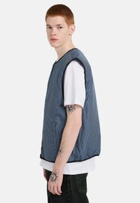 Gilet a quadretti blu e bianchi realizzato in tessuto leggero, con scollo rotondo, design senza maniche e rifiniture nere lungo i bordi.