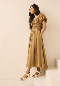 Robe longue marron avec manches bouffantes et corsage smocké ; associée à des sandales compensées dorées. Fond avec un mur texturé beige clair.