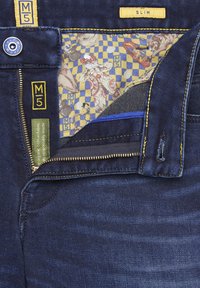 Jean en denim slim fit avec un tissu bleu foncé, des coutures et des accents jaunes, un zip, et une doublure intérieure à motif à carreaux.