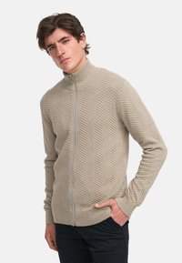 Suéter beige de punto con cremallera, cuello acanalado, patrón de espiga y dos bolsillos laterales. Textura suave y diseño ajustado.