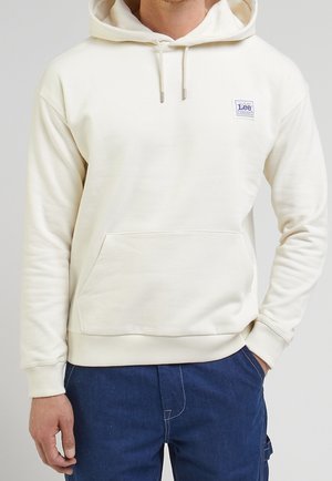 Muž v krémové mikině s kapucí a přední kapsou, na které je nášivka Lee Workwear, spárovaný s modrými džíny, s rukou v kapse.