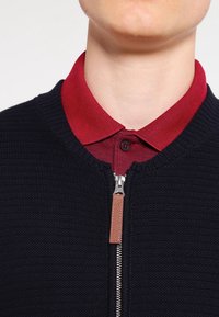 Marinblå stickad zip-up tröja med texturerad finish, i kombination med en röd skjorta med krage som har knapppdetaljer och kontrasterande tyger.