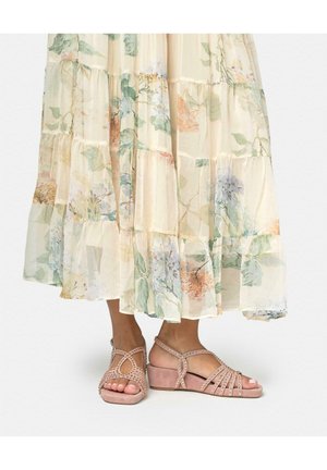 Pies con sandalias rosas con tachuelas y tacón bajo, combinadas con una falda larga floral en color crema y fluida.