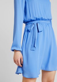 Blå, knälång klänning med långa, lätt utsvängda ärmar. Har en rynkad midja och ett knytbälte. Slät tyg med en mjuk lyster.