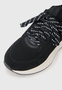 Black athletic shoe featuring a combination of mesh and suede materials, textured surface, thick white midsole, and woven black-and-white laces.