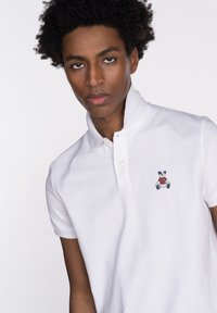 Camisa polo blanca de algodón con cuello corto, que presenta un logo bordado de un oso de peluche en azul y rojo en la parte izquierda del pecho.