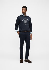 Hombre que lleva una sudadera azul marino con el texto "GANT Sportswear New Haven 1949", camisa azul claro, pantalones azul marino, cinturón marrón y mocasines marrón oscuro, de pie.