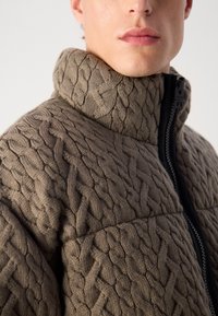 Brun strikket sweater med høj krave og sort lynlås. Har et tekstureret kabelstrik mønster og en tætsiddende design.