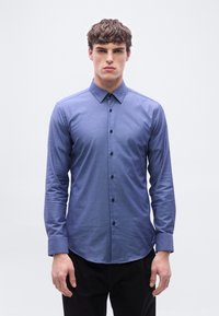 Νέος άνδρας που στέκεται, φορώντας στενή μπλε πουκάμισο με κουμπιά μακρυμάνικο και μαύρο παντελόνι, μπροστά σε απλό φωτεινό φόντο.