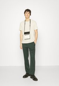 Beige kortærmet t-shirt, mørkegrønne bukser og sorte sko. En lille sort og hvid crossbody-taske bæres om halsen.