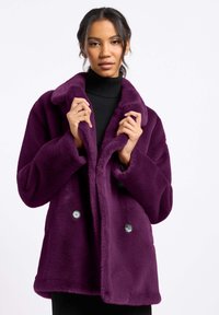 Fioletowy płaszcz z sztucznego futra z szerokim kołnierzem, podwójnym zapięciem i dużymi guzikami. Miękka tekstura i luźny krój odpowiedni do noszenia na warstwy.