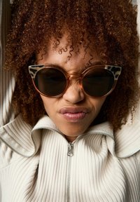 Lunettes de soleil en forme de yeux de chat avec une monture brune translucide et des accents imprimés animaliers contrastants. Portées avec une veste zippée légère à côtes.