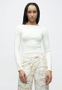 Bluză albă cu mâneci lungi, cu textură rib și design în formă de V, cu manșete cu nasturi, asortată cu pantaloni deschiși la culoare, cu modele cu motive de lanțuri aurii.