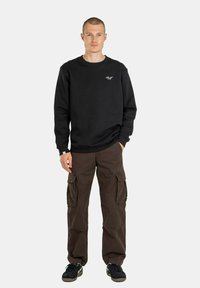 Schwarzer Sweatshirt mit gerippten Bündchen und Ausschnitt, versehen mit einem kleinen gestickten Logo. Kombiniert mit braunen Cargo-Hosen mit seitlichen Taschen.