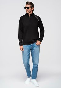 Sudadera negra con cremallera y cuello, que presenta un acento de texto blanco en el hombro y puños de canalé, combinada con jeans azules y zapatillas blancas.