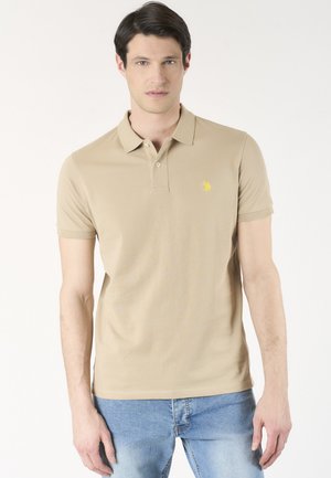Giovane uomo che indossa una polo beige a maniche corte con logo giallo e jeans azzurri, in piedi davanti a uno sfondo bianco semplice.